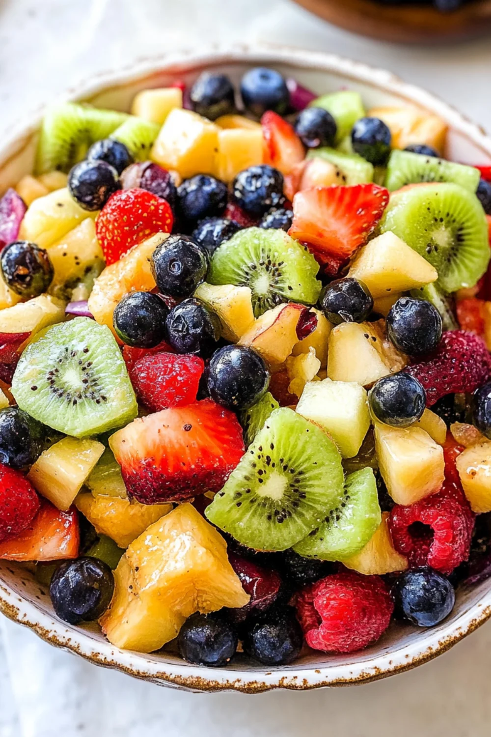 A vibrant and fresh bowl of mixed fruit salad featuring strawberries, blueberries, kiwi, pineapple, and raspberries, arranged neatly and topped with a sprinkle of mint.