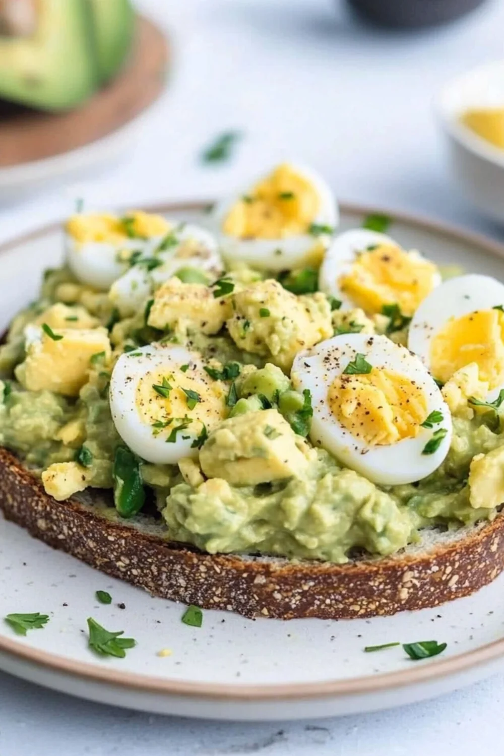 A close-up of Avocado Egg Salad on toasted bread. The sandwich is topped with halved hard-boiled eggs, fresh parsley, and a sprinkle of black pepper, showcasing a creamy avocado and egg mixture.