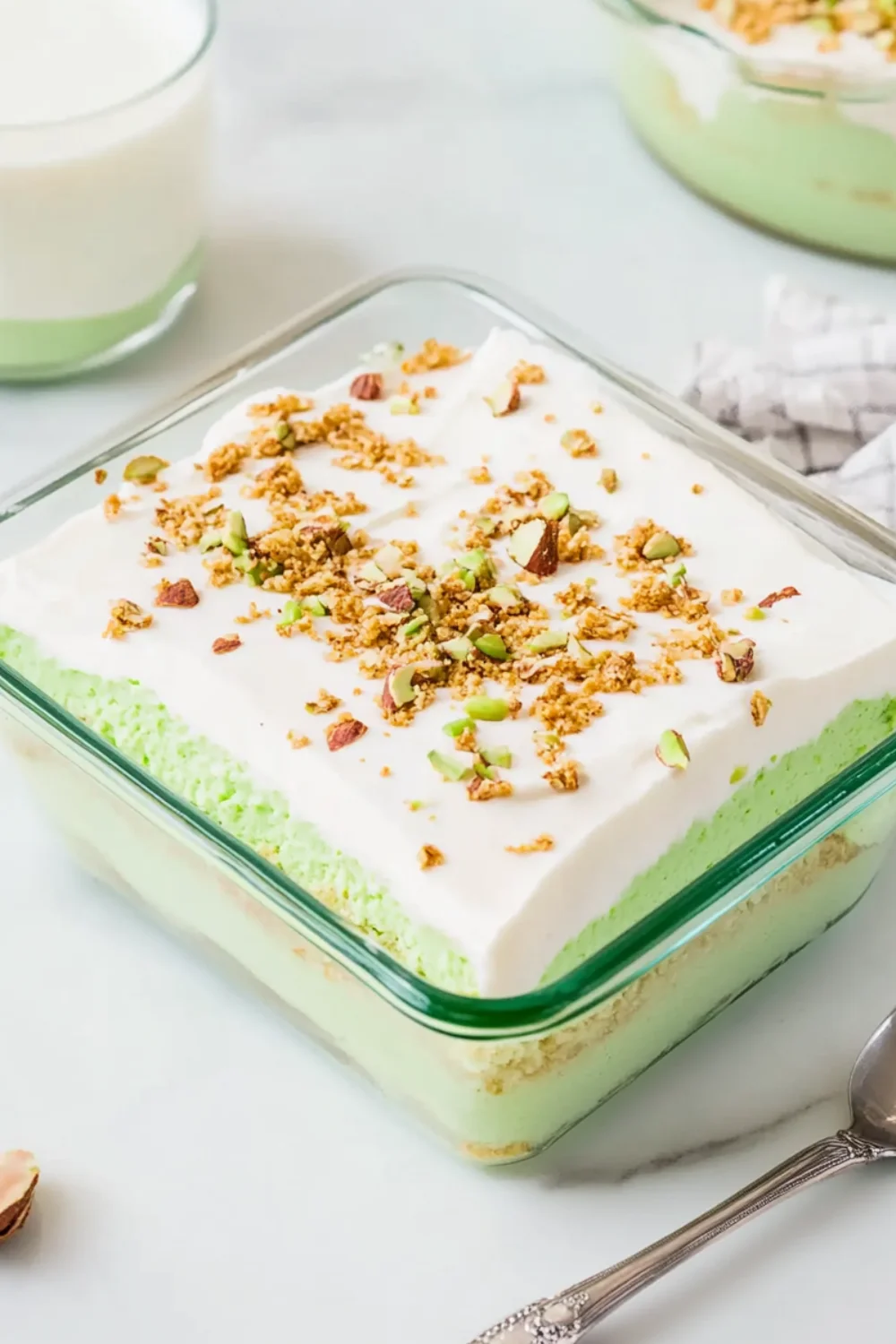 Layered pistachio pudding dessert topped with whipped cream and chopped nuts in a glass dish.
