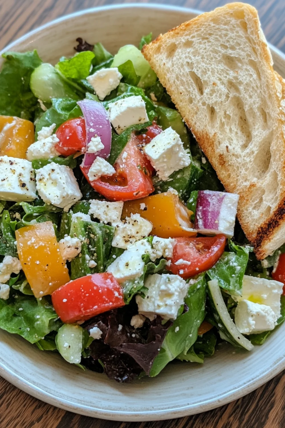 A fresh Greek salad with tomatoes, cucumbers, red onion, feta cheese, and mixed greens, served with a slice of toasted bread.