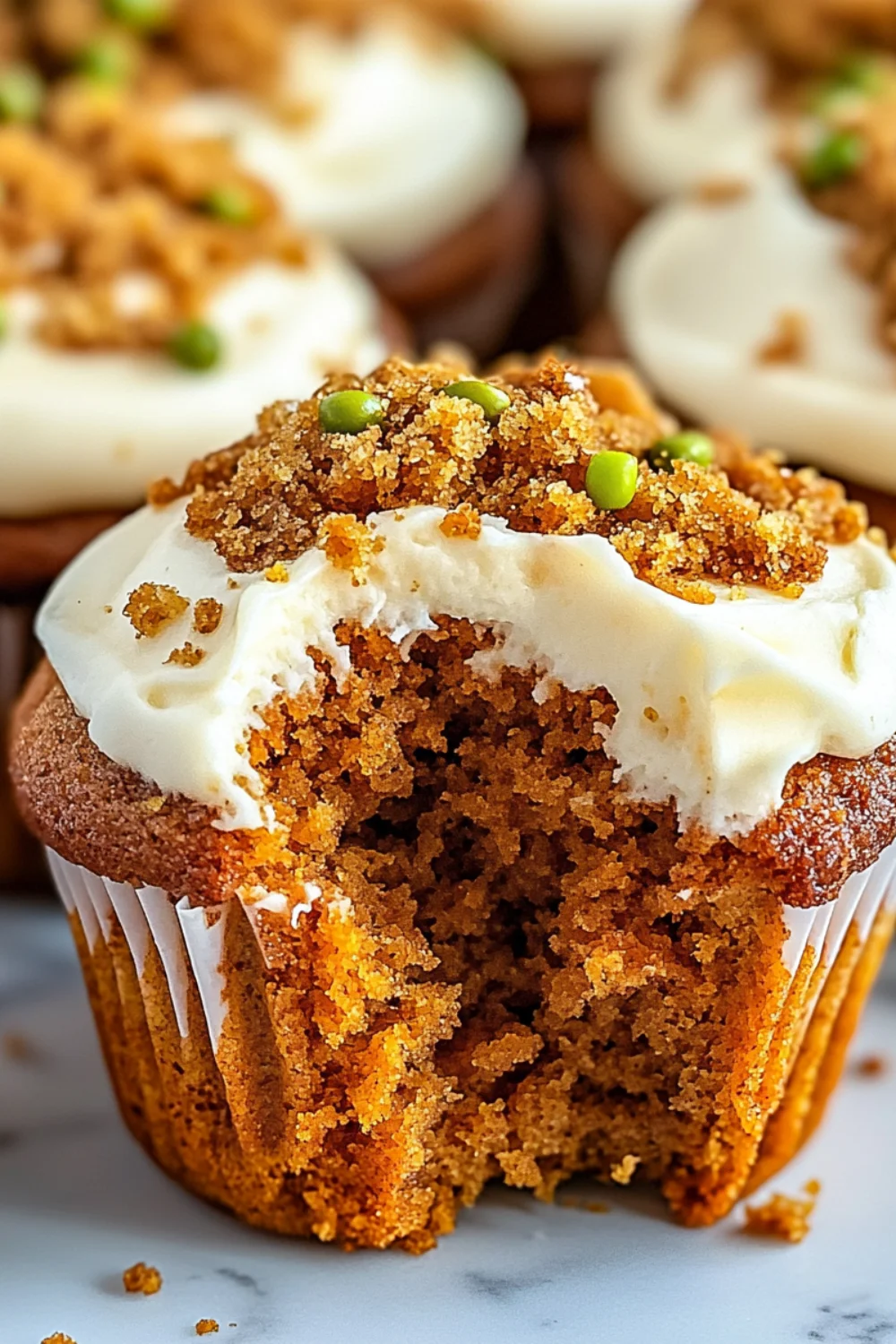 A freshly baked carrot cake muffin with a bite taken out, topped with cream cheese frosting and crumbles.